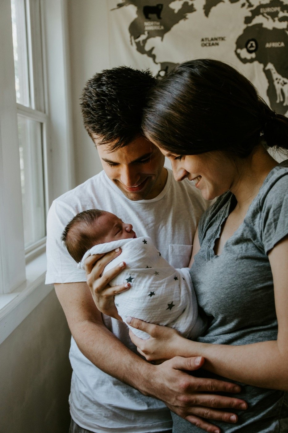 Parents smiling down at their wrapped newborn beside a window